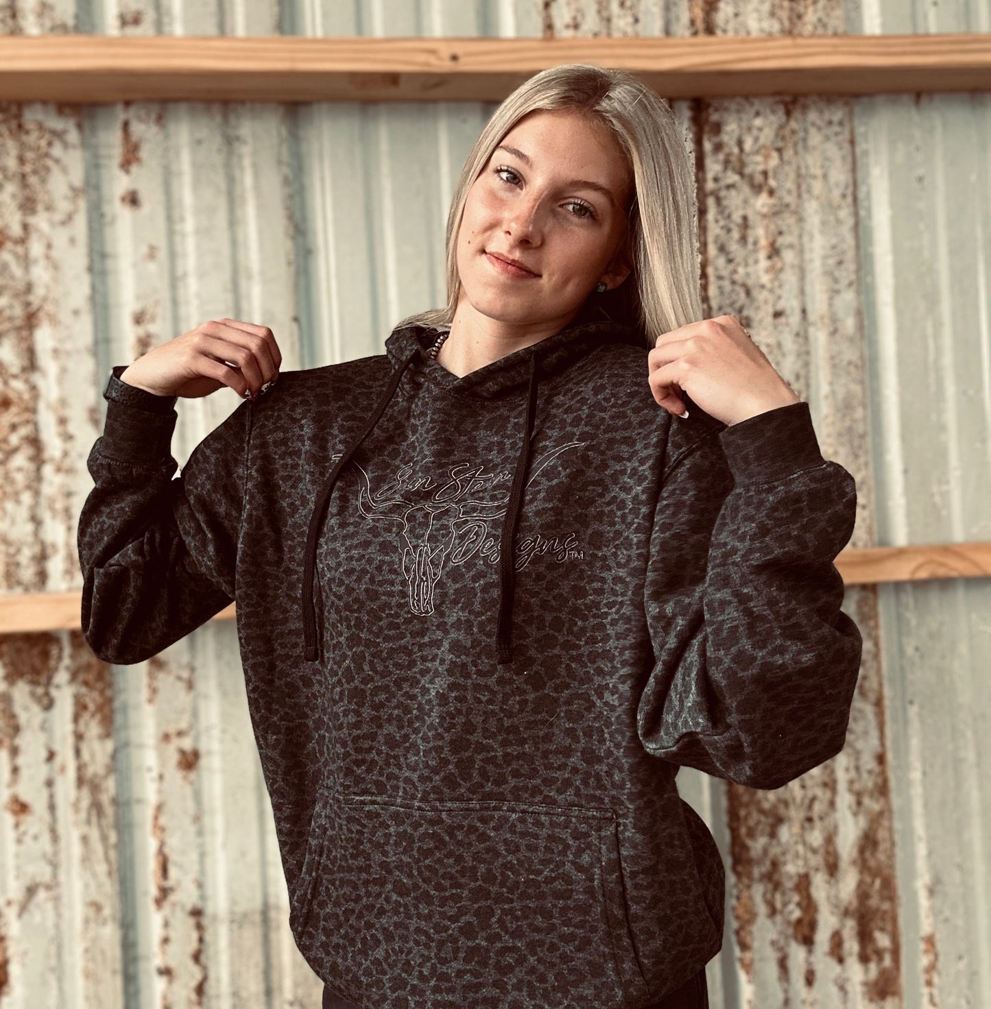 Person wearing a dark patterned hoodie with text, standing against a rustic metal wall.