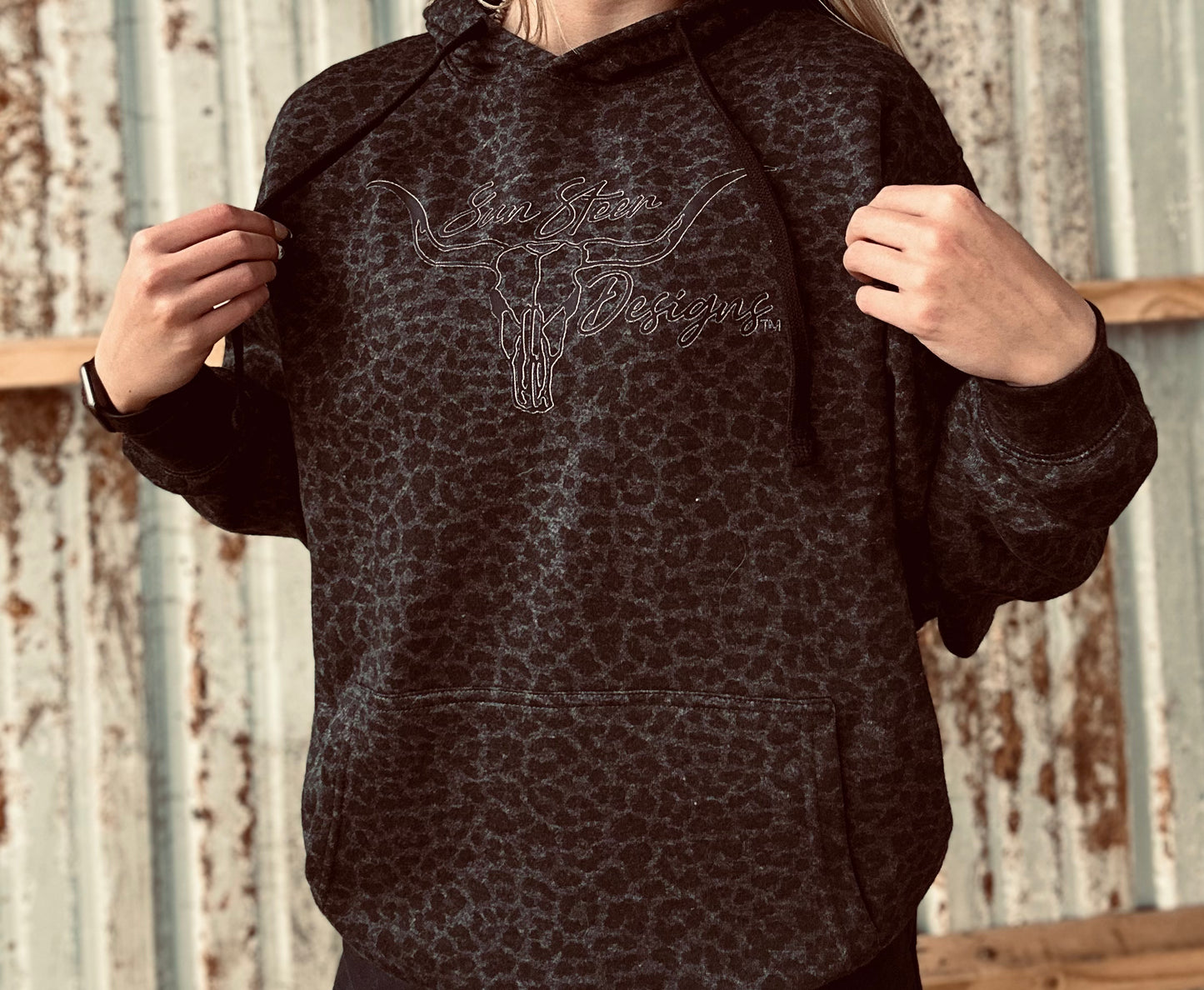 Person wearing a dark patterned hoodie with branding against a rustic metal wall.