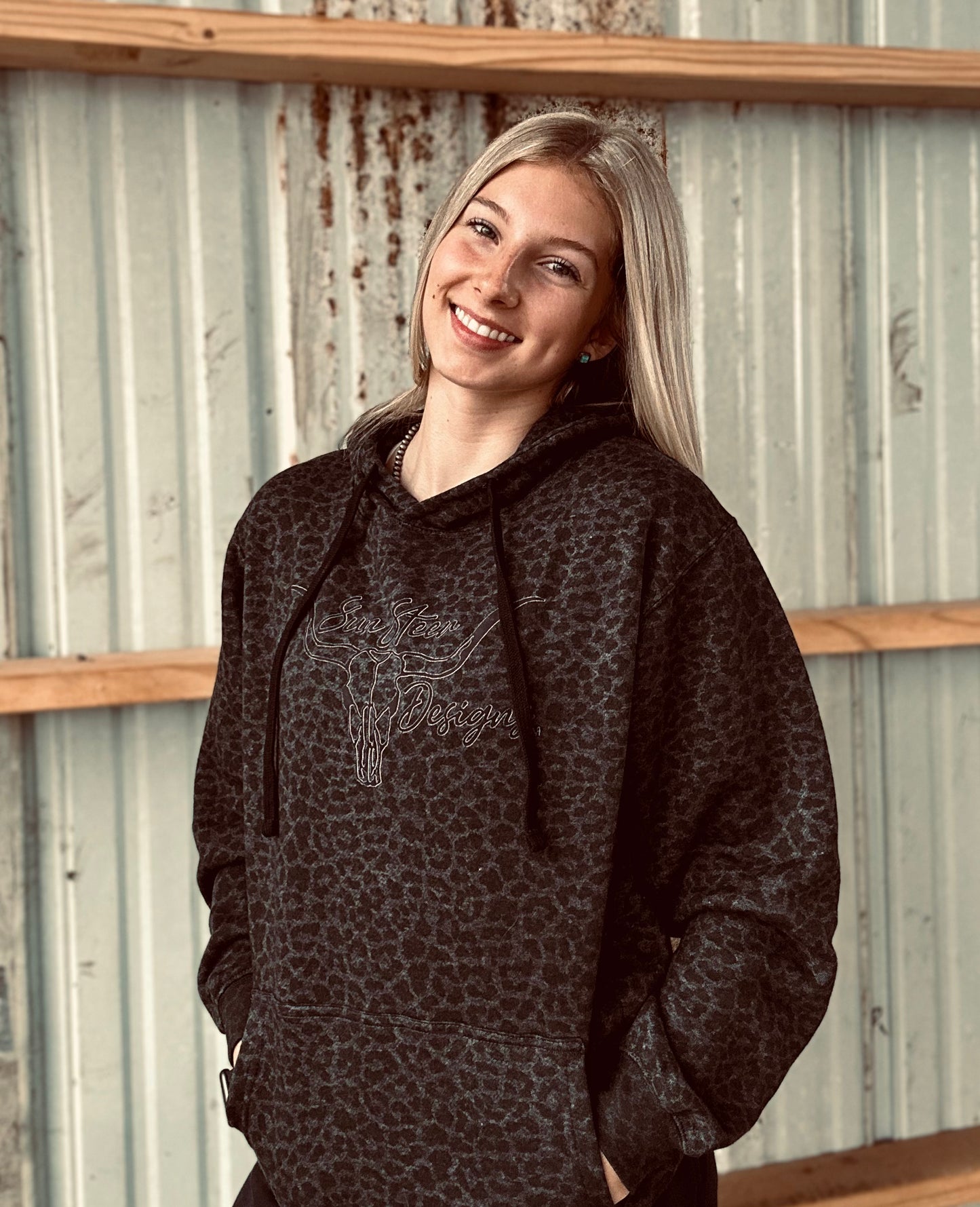 Woman wearing a dark hoodie with a pattern against a corrugated metal wall.