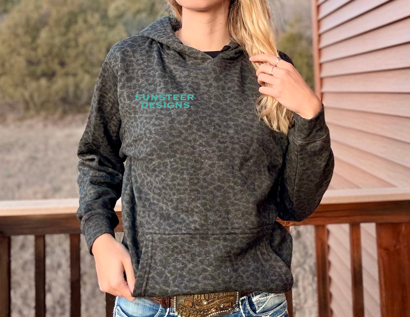 Person wearing a black leopard hoodie with teal 'Sunsteer Designs' logo, standing on a wooden deck.