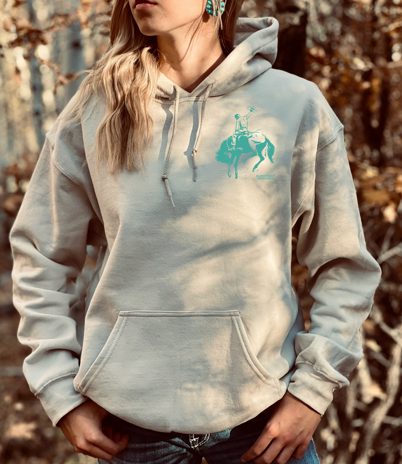 Person wearing a tan hoodie with a turquoise graphic of a bucking bronc design outdoors and rider in an outline of cards
