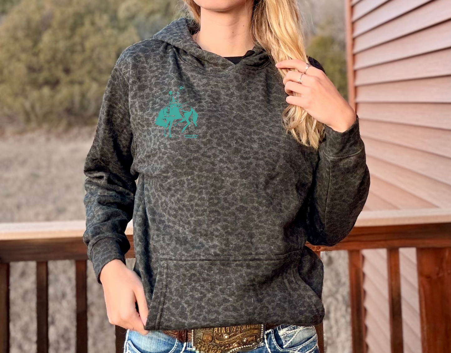 Person wearing a black leopard hoodie with a bucking bronc logo left chest, standing on a wooden deck.