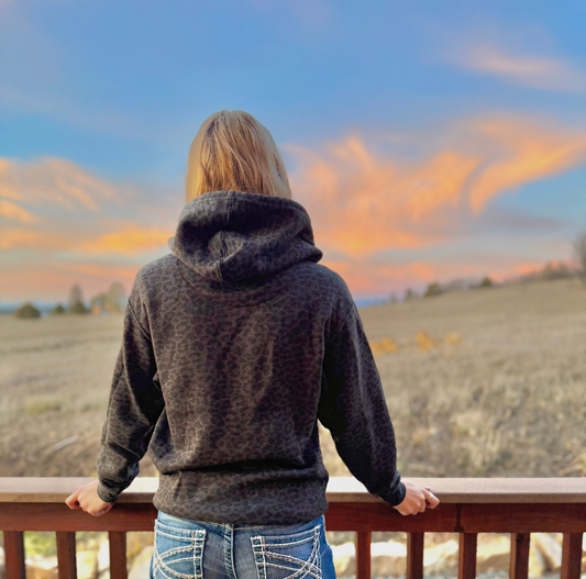 Person wearing a black leopard hoodie looking at a sunset over a field from a balcony.