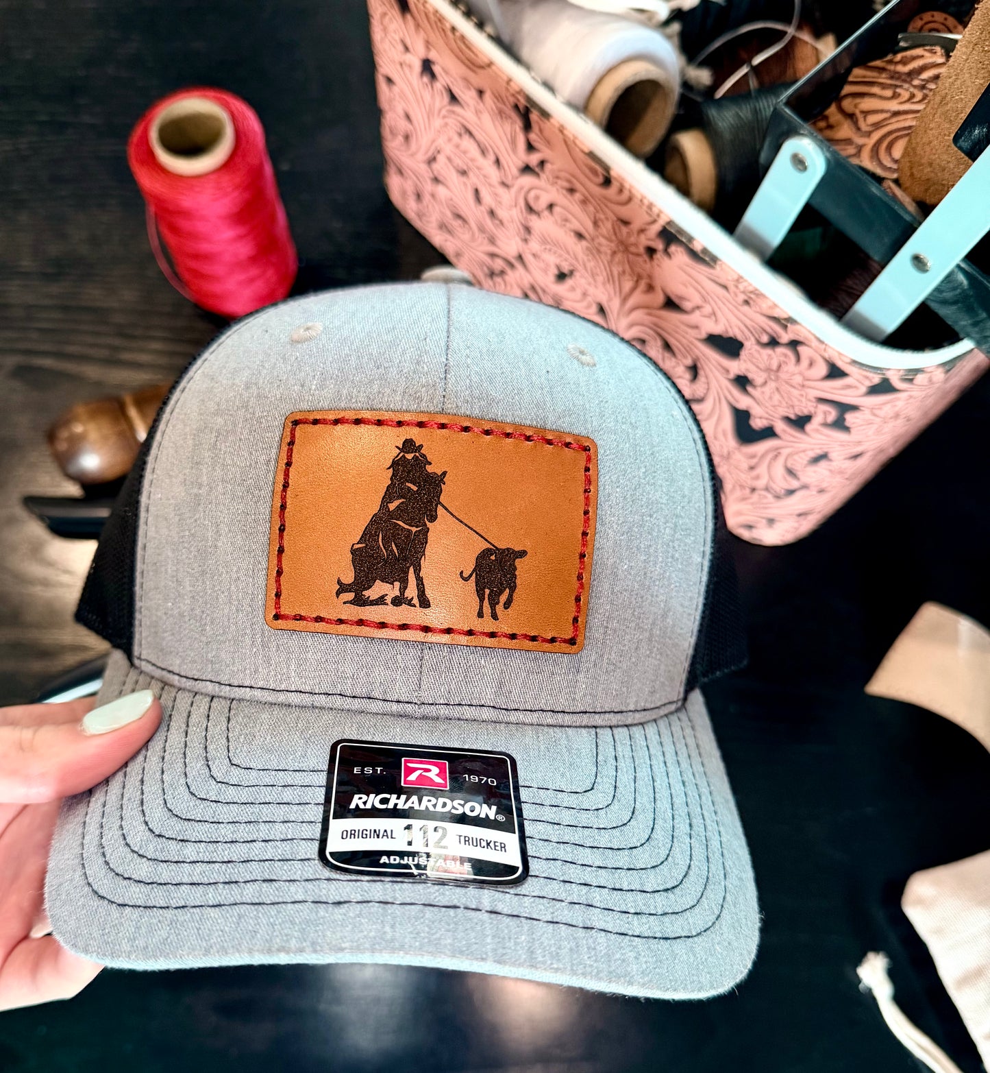 Gray cap with leather patch featuring a breakaway roper design, held by a hand on a dark surface.