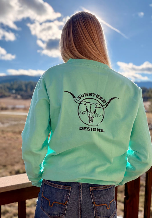 Person wearing a light green sweatshirt with 'Sunsteer Designs' logo, standing outdoors with a scenic background.