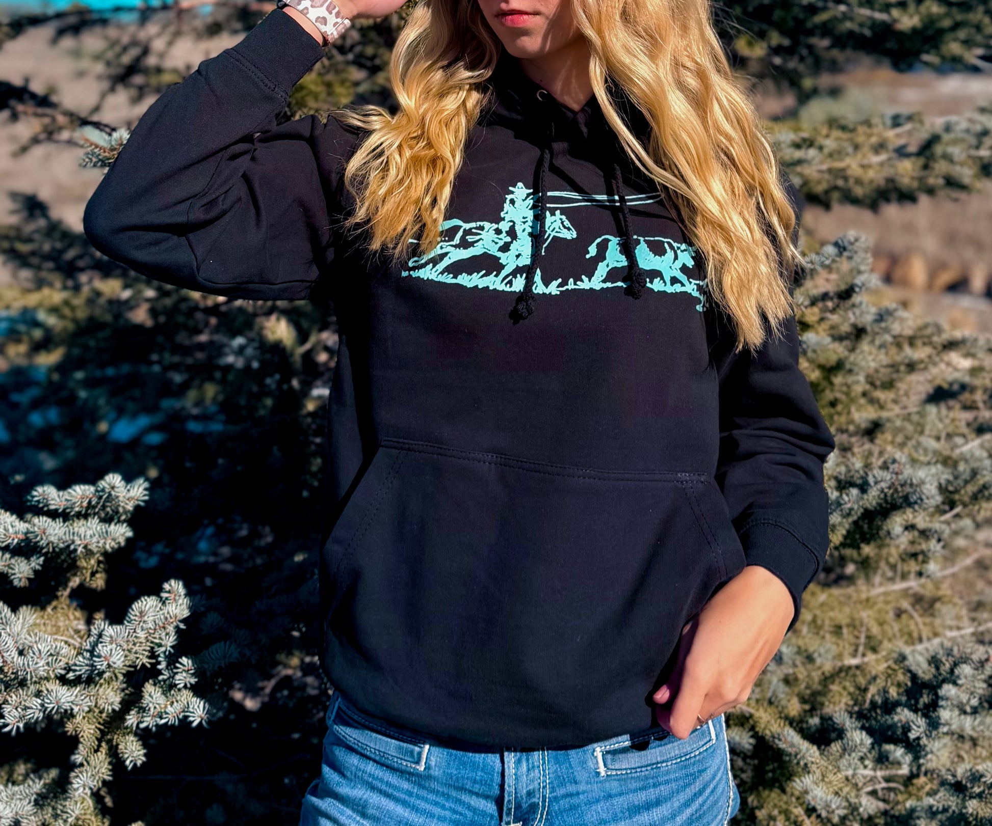Black hoodie with a teal roping design worn by a girl standing in front of a tree. 