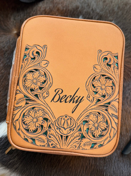 A tan leather zippered case with floral cutout detailing and the name 'Becky' printed on the front, placed on a cowhide surface.