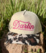 A beige baseball cap with bright pink embroidered script reading 'Darlin' on the front, resting on a rock outdoors with green grass in the background. The brim has a brown, black, and beige cow-print pattern.