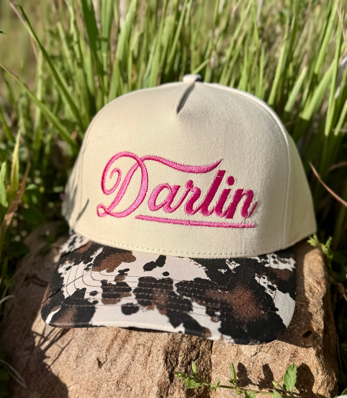A beige baseball cap with bright pink embroidered script reading 'Darlin' on the front, resting on a rock outdoors with green grass in the background. The brim has a brown, black, and beige cow-print pattern.