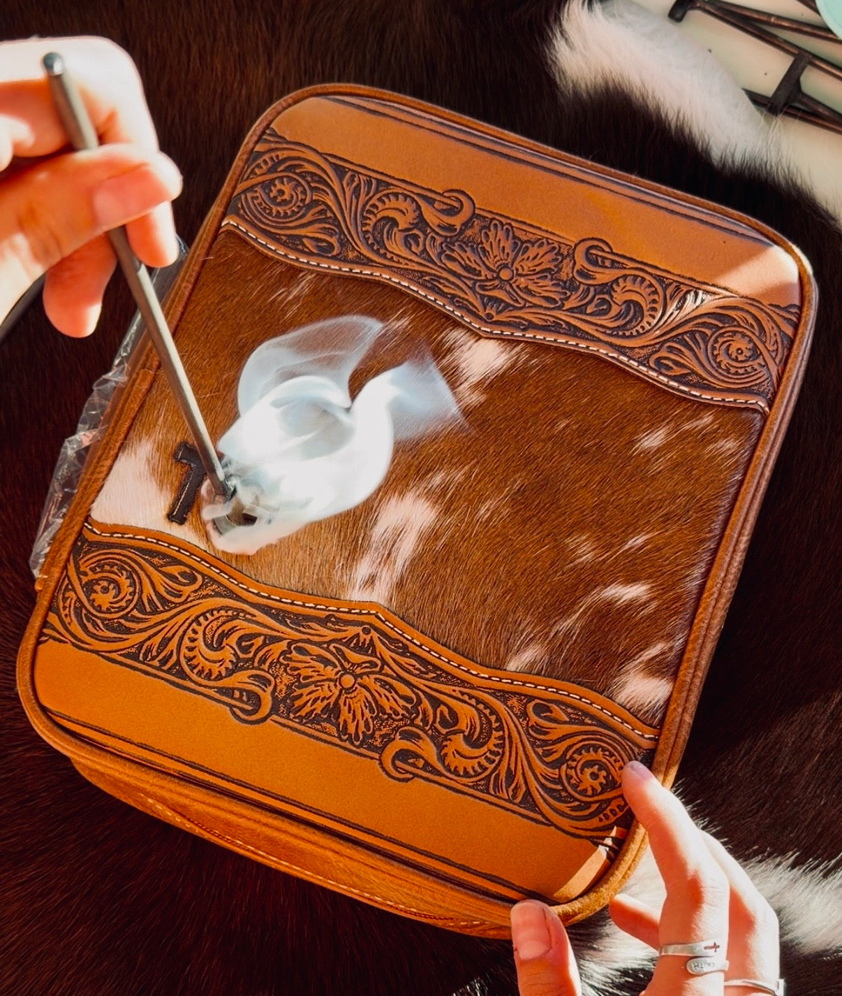 A brown leather and cowhide zippered case with ornate floral tooling, held open by two hands. A metal tool is being used to apply a white adhesive or seal to the front flap.