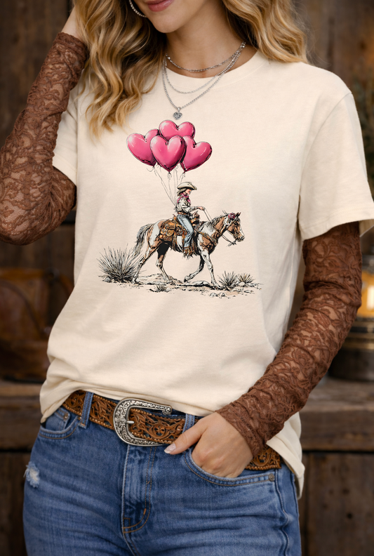 Person wearing a white t-shirt with a graphic of a person on a horse with pink balloons, paired with brown lace sleeves and blue jeans.