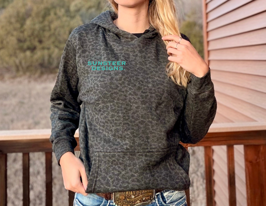 Person wearing a black leopard hoodie with teal 'Sunsteer Designs' logo, standing on a wooden deck.