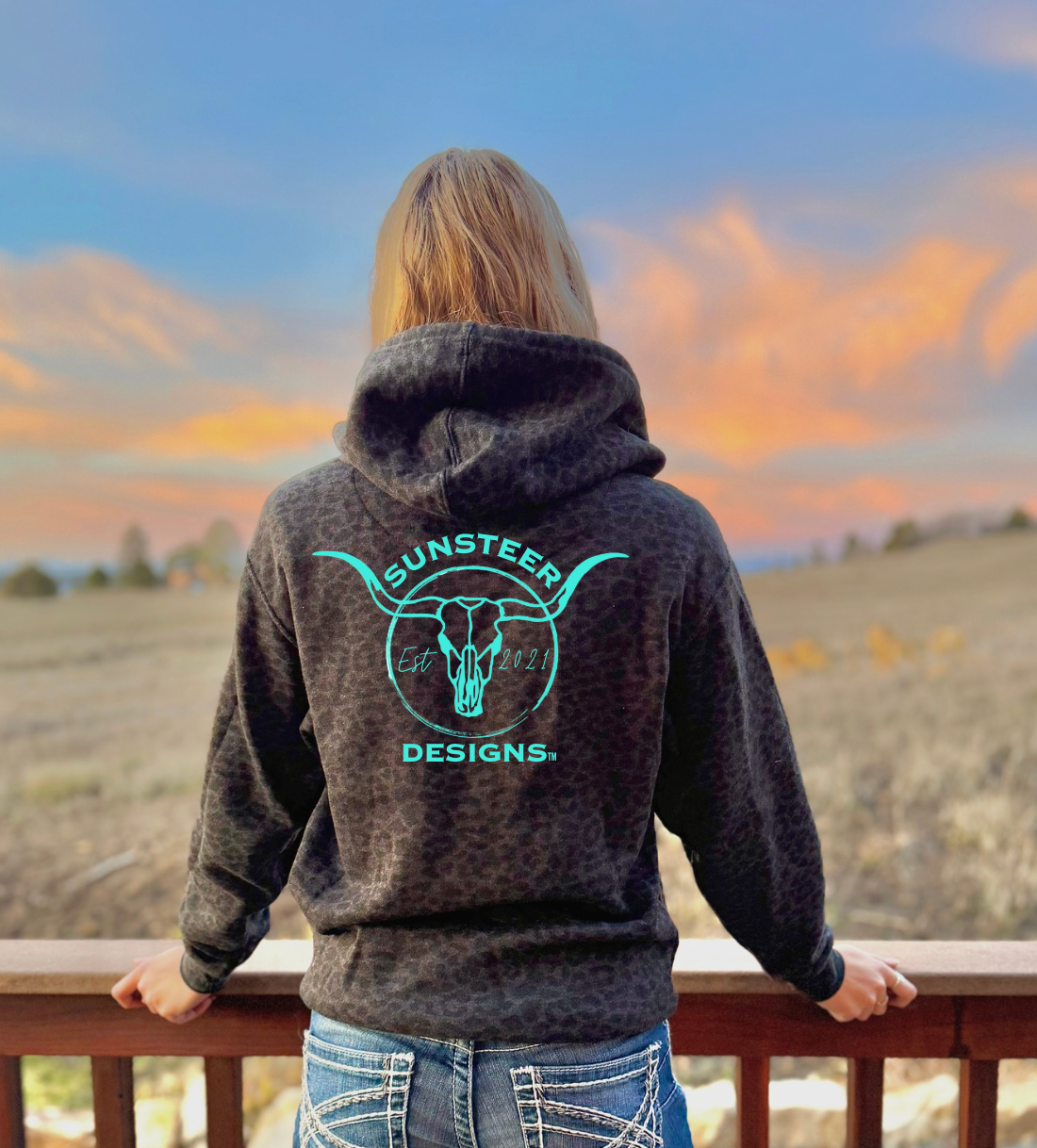 Person wearing a hoodie with 'Sunsteer Designs' logo, standing on a balcony overlooking a scenic landscape.