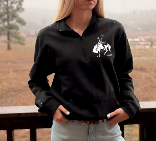 Person wearing a black 1/4 zip sweatshirt with a white graphic of a bucking bronc horse and rider, standing outdoors.