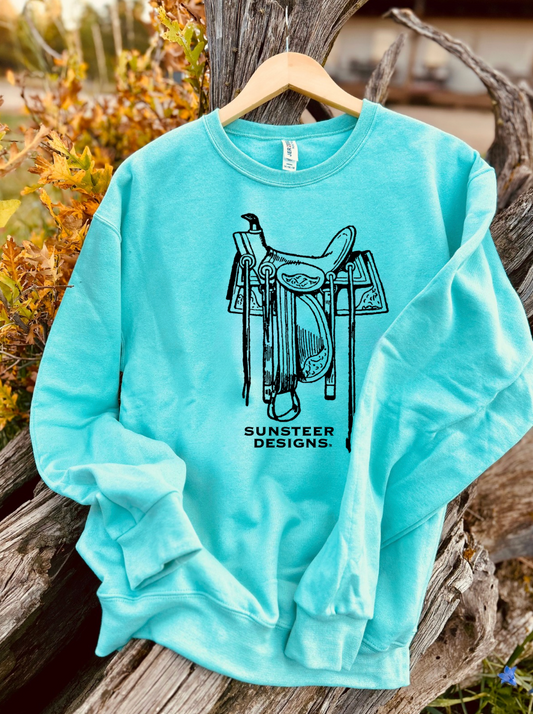 Mint sweatshirt with a saddle graphic and 'Sunsteer Designs' text on a wooden surface.