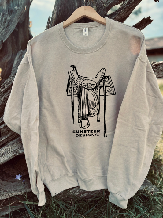 Tan sweatshirt with a saddle graphic and 'Sunsteer Designs' text on a wooden background