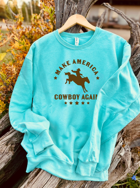 Mint sweatshirt with brown 'Make America Cowboy Again' text and graphic on a wooden surface.
