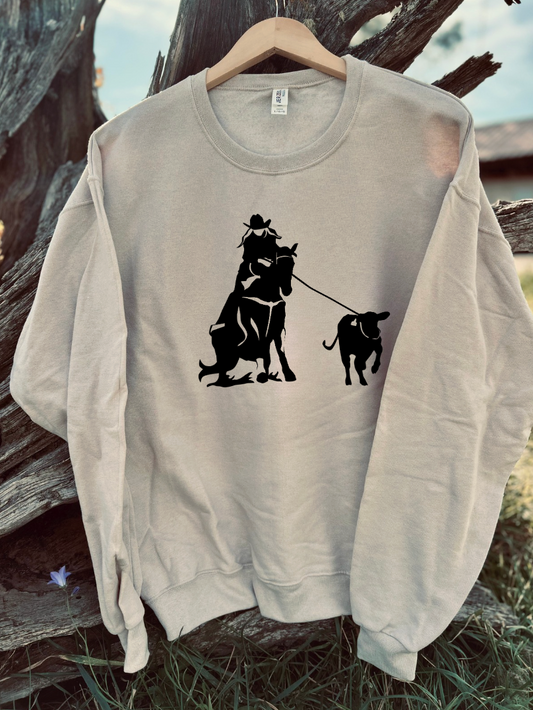 Sandstone sweatshirt with black graphic of a person riding a horse and roping a breakaway calf, on a hanger against a natural background.