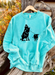 Mint sweatshirt with black graphic of a person riding a horse and roping a breakaway calf, on a hanger against a natural background.
