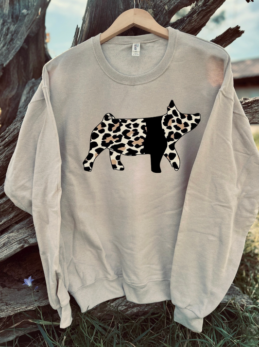 Tan sweatshirt with a leopard print cow design hanging on a wooden hanger against a natural background.