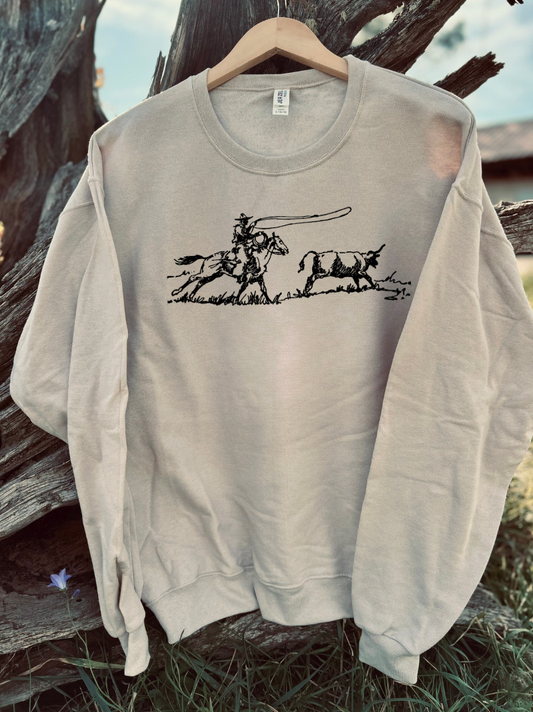 Sandstone sweatshirt with a graphic design of a cowboy roping a steer in a pasture outline, on a wooden surface