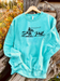 Mint sweatshirt with a graphic design of a cowboy roping a steer in a pasture outline, on a wooden surface