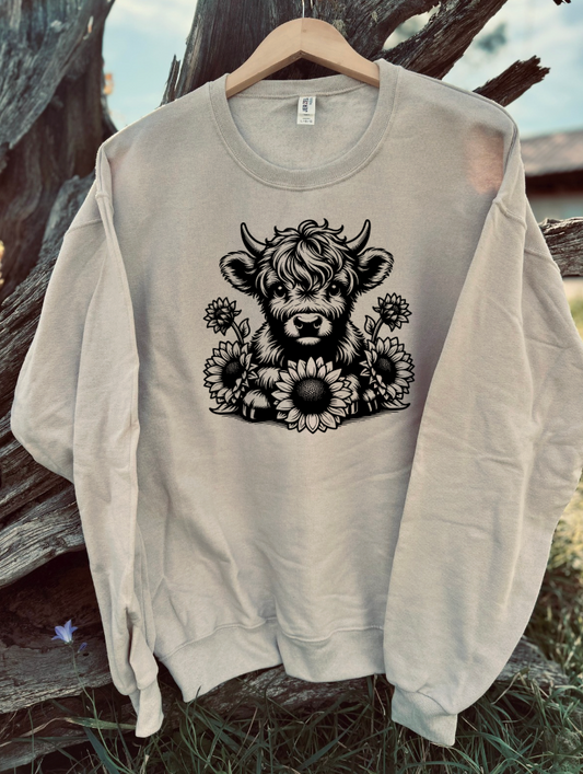 Tan sweatshirt with a black design of a highland cow and sunglowers flowers on a wooden background.