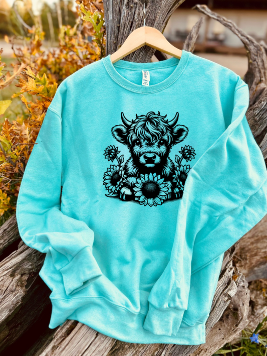 Mint sweatshirt with a black outline of a highland cow animal and sunflower design hanging on a stump