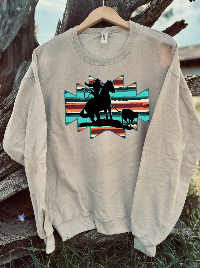 Tan sweatshirt with a graphic of a silhouette of a person on horseback roping a steer against a colorful aztec background, hanging on a wooden surface.