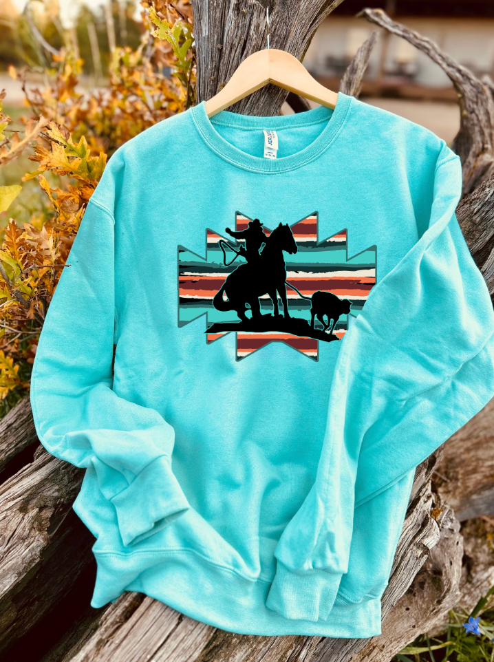 mint sweatshirt with a graphic of a horse and rider roping a breakaway steer with an aztec background on a wooden surface.