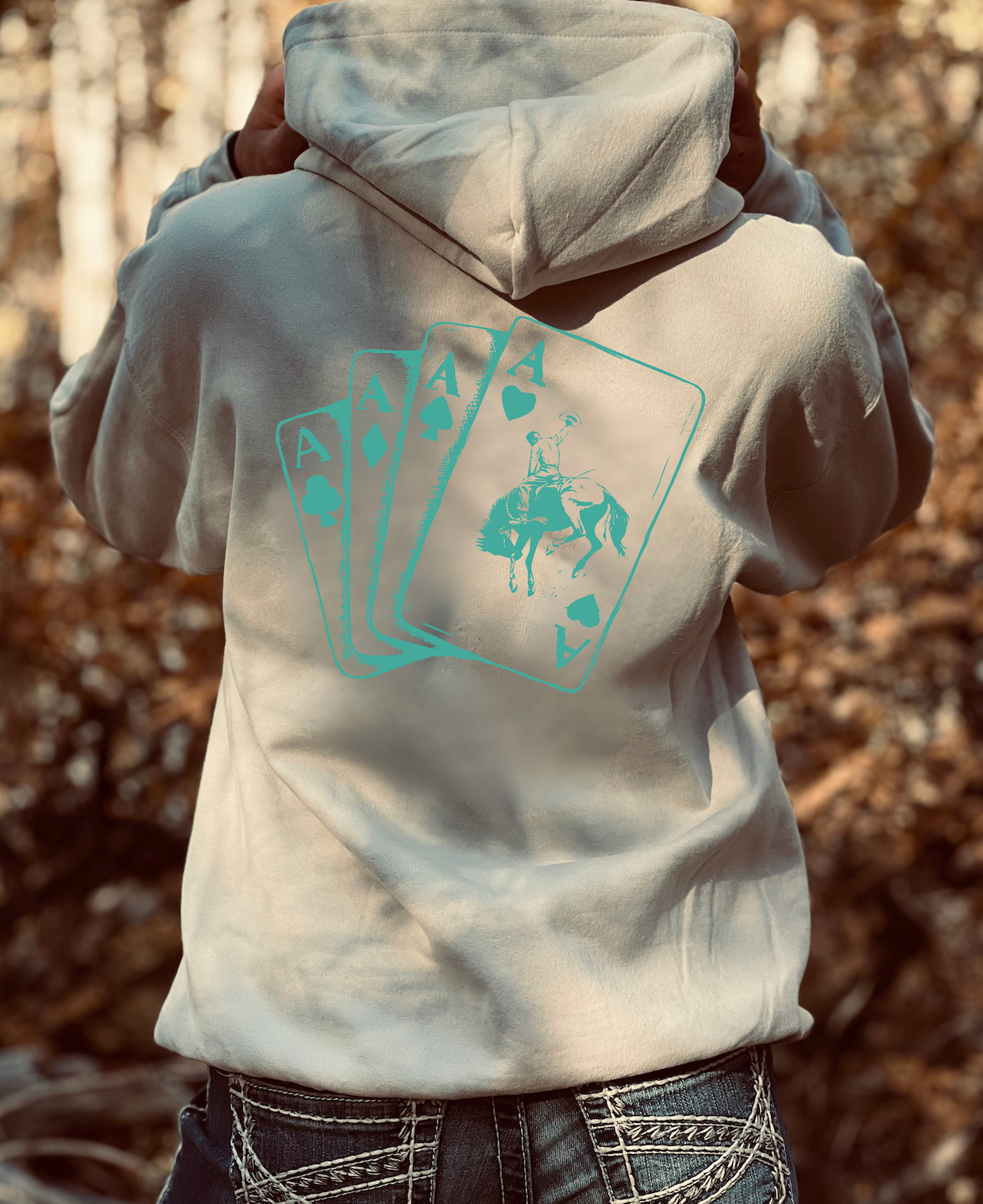 Person wearing a tan hoodie with a turquoise graphic of a bucking bronc design outdoors and rider in an outline of cards 