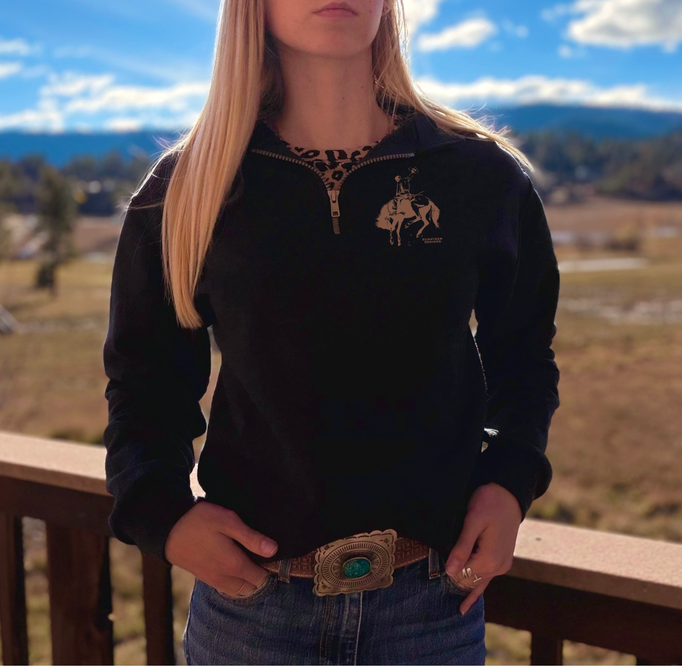 Person wearing a black 1/4 zip hoodie with  a brown bucking bronc and rider outline, standing outdoors with mountains in the background